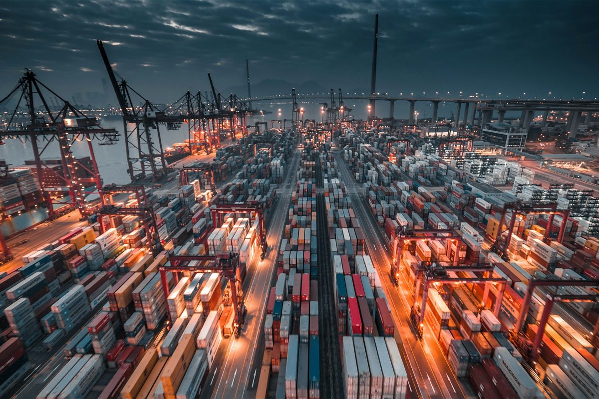 A container port at dusk, with cranes and long rows of stacked shipping containers lit in orange