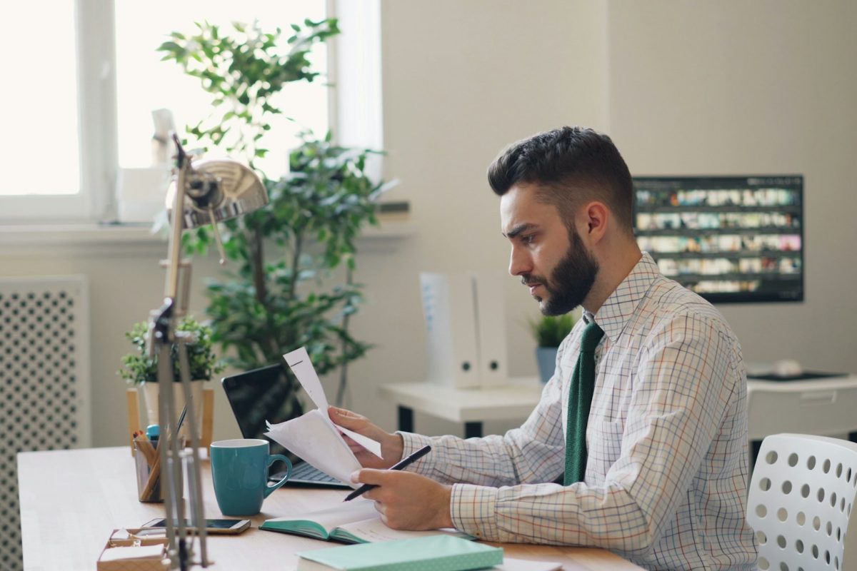 A man in a shirt and tie reviewing papers at a desk in a bright office