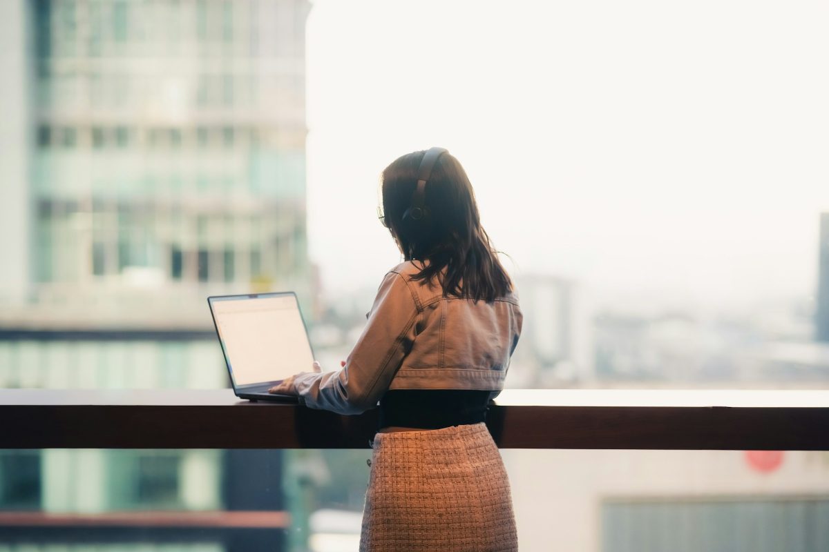 Frau mit Kopfhörern arbeitet am Laptop an einem Fenstertresen mit Blick über die Stadt