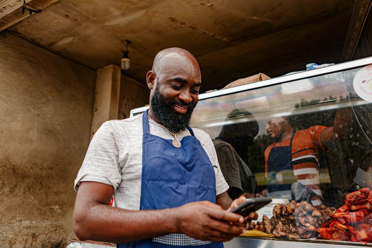 A smiling stall owner in a blue apron checking his phone at his counter