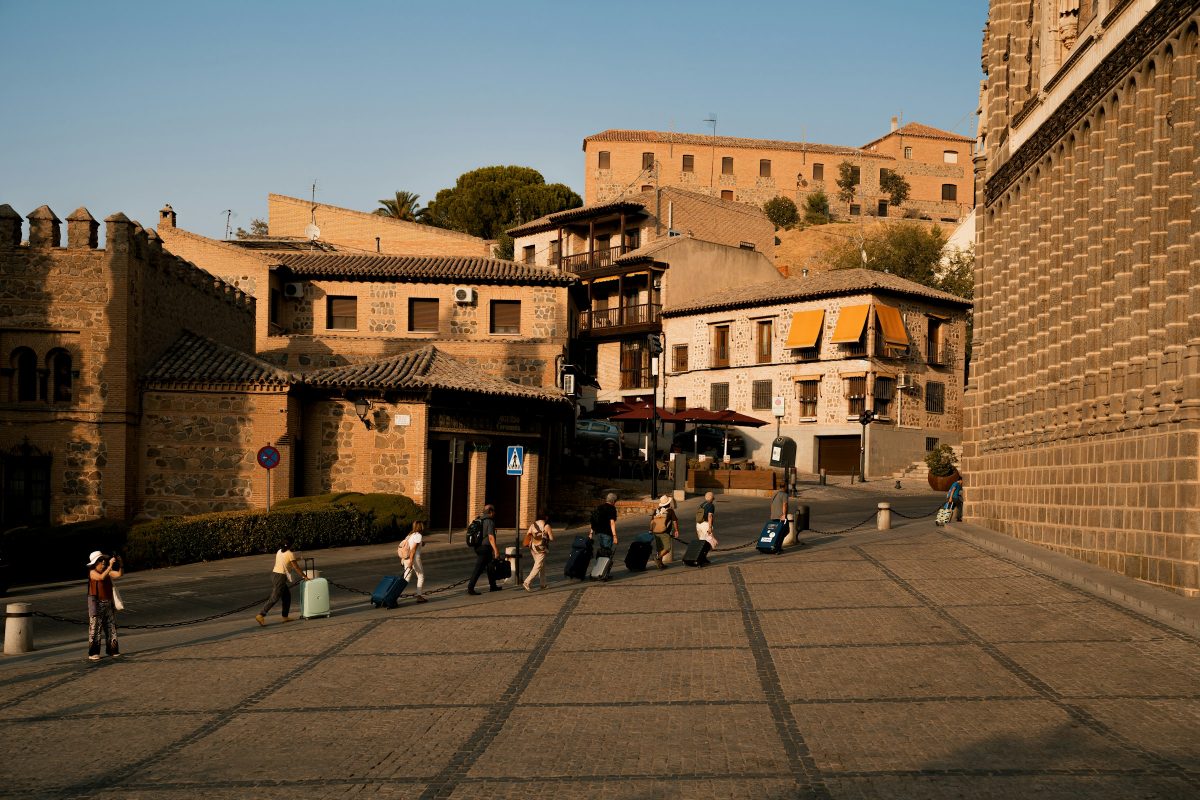 Viajeros con maletas cruzando una plaza empedrada y soleada de una ciudad europea antigua
